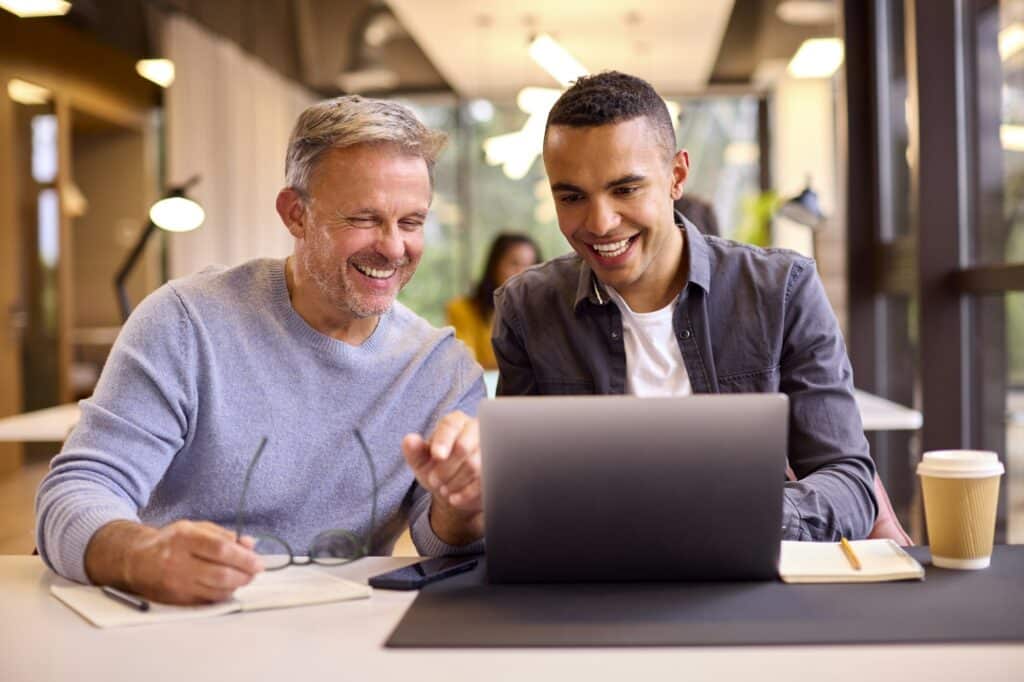 Mature Businessman Mentoring Younger Colleague Working On Laptop At Desk